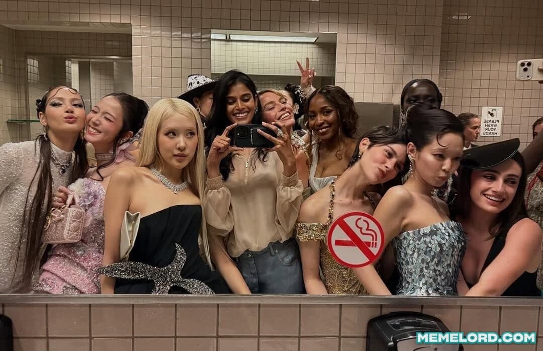 A tightly packed group of glamorous guests poses for a bathroom mirror selfie at the Met Gala. One person holds a phone at the center while the group fills the frame; tiled walls, a no-smoking sign, and a bottom-right MEMELORD.COM watermark are visible.