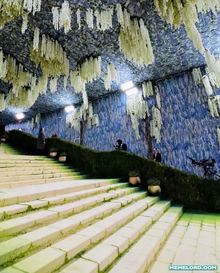 Vertical photograph of a broad pale stone staircase rising toward the upper left beneath a dense canopy of hanging white and pale green flowers. Clipped hedges border the stairs, photographers and event staff stand in the distance, blue floral walls surround the space, green lighting washes over the steps, and a MEMEL은