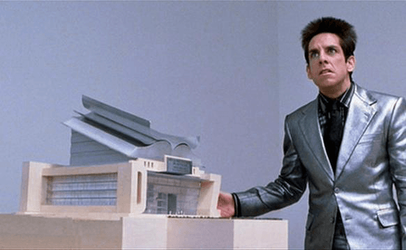 Derek Zoolander in a shiny silver suit gestures toward a small architectural model displayed on a table against a plain gray background.