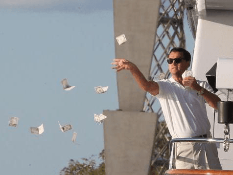 A man wearing sunglasses and a white shirt stands on a yacht and throws several banknotes into the air beside him.