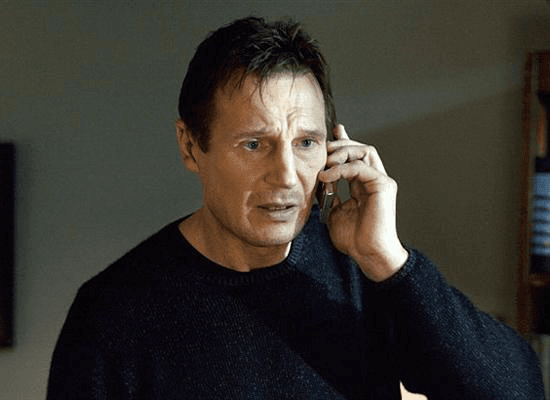 A close-up film still of Liam Neeson holding a phone to his ear, looking tense and intensely focused while wearing a dark sweater.