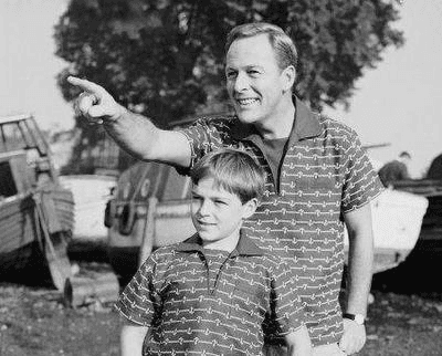 A smiling father in a patterned shirt stands behind a young boy in a matching shirt and points toward something off the left side of the image.