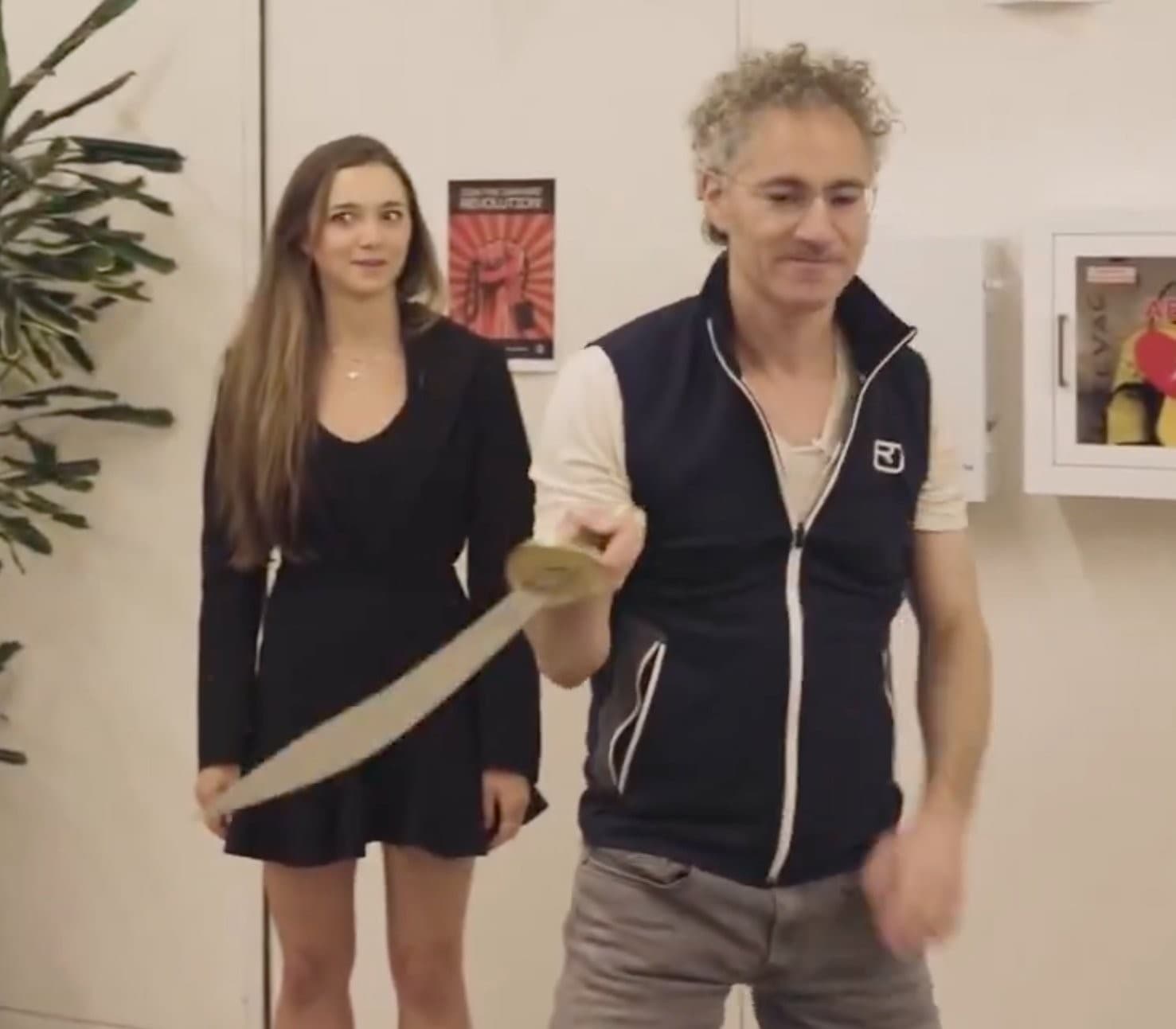 A gray-haired man in a dark vest stands in an office-like room while holding a large pale sword-shaped prop across the foreground; a woman in a black dress stands behind him.