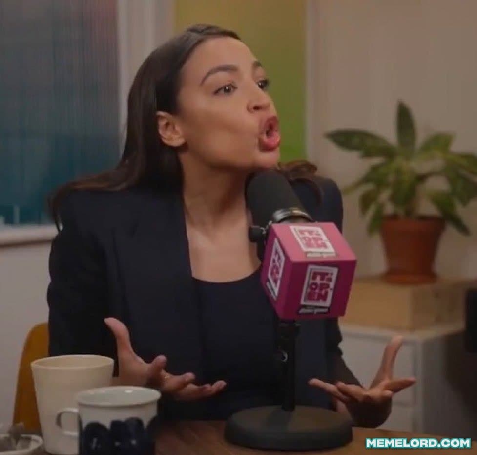Alexandria Ocasio-Cortez sits at a table speaking into a branded microphone with both hands raised and open, expressive gestures. A mug and a potted plant are visible in the background.