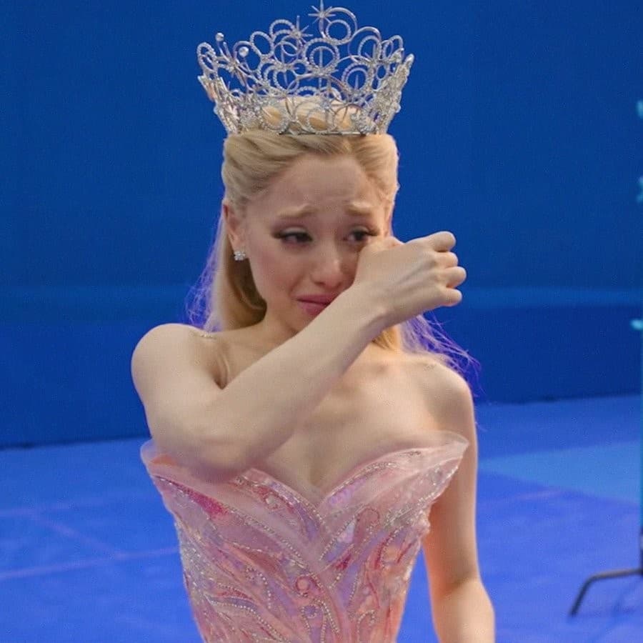 Ariana Grande in character as Glinda wears a sparkling tiara and pink gown while wiping tears from her face with her arm against a blue background.