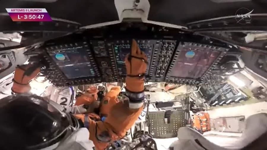 A wide-angle camera view inside the Orion spacecraft shows astronauts in orange flight suits beneath a bank of cockpit screens and controls, with the cabin appearing rotated upside down. A launch countdown graphic is at the upper left and a NASA logo is at the upper right.