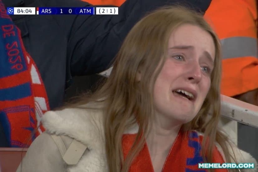 A young female football supporter wearing a red-and-white scarf cries in the stands, with a broadcast scoreboard showing Arsenal leading Atlético Madrid 1–0 at 84:04 and a small Memelord.com watermark in the lower-right corner.