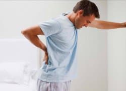 A young man in a light blue shirt and pale shorts bends forward in pain, pressing one hand against his lower back while extending the other arm toward a light-colored wall.