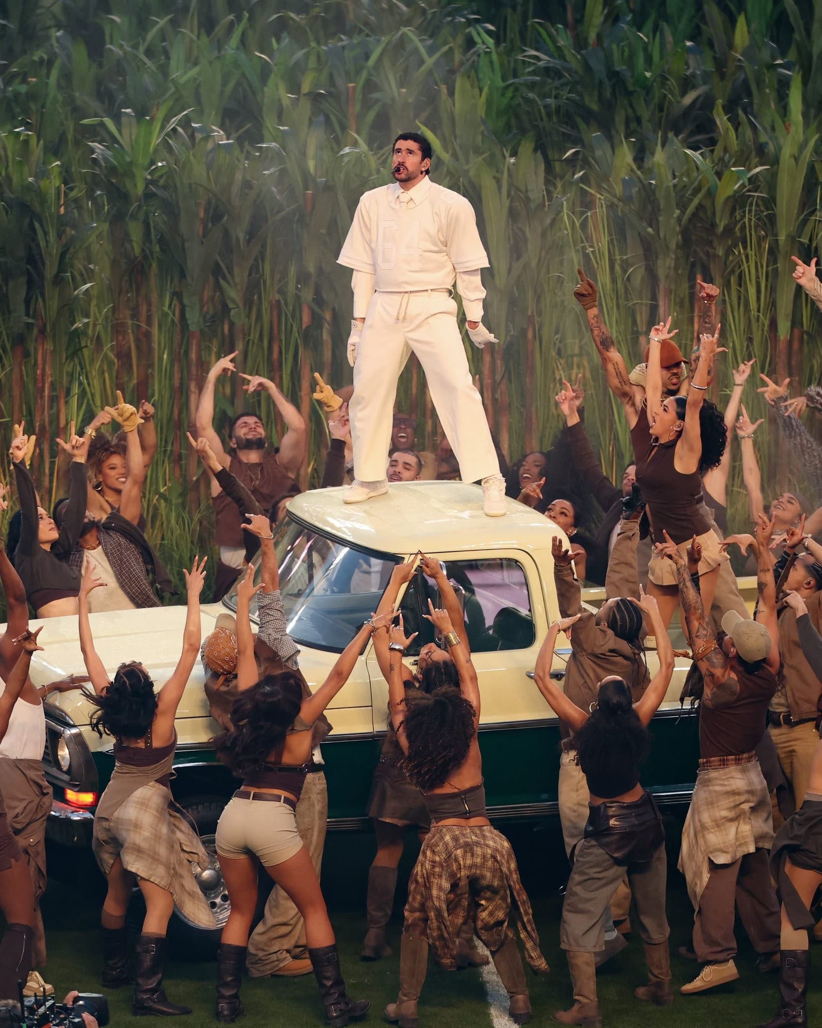 A performer in an all-white outfit stands on the roof of a cream-colored vintage car, surrounded by a large cheering crowd in front of tall green plants.