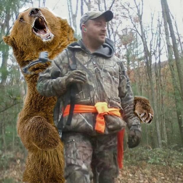 A camouflaged hunter walks through leafless woods while a large roaring bear stands directly behind him with its paws raised.