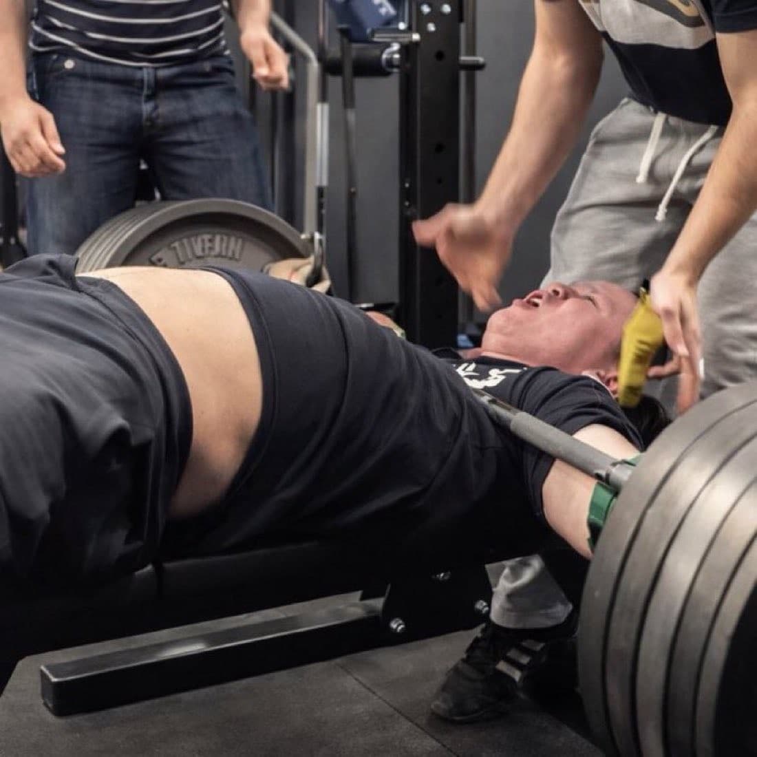 A man lies on a gym bench beneath a heavily loaded barbell, grimacing as several people reach toward him to help with the lift.
