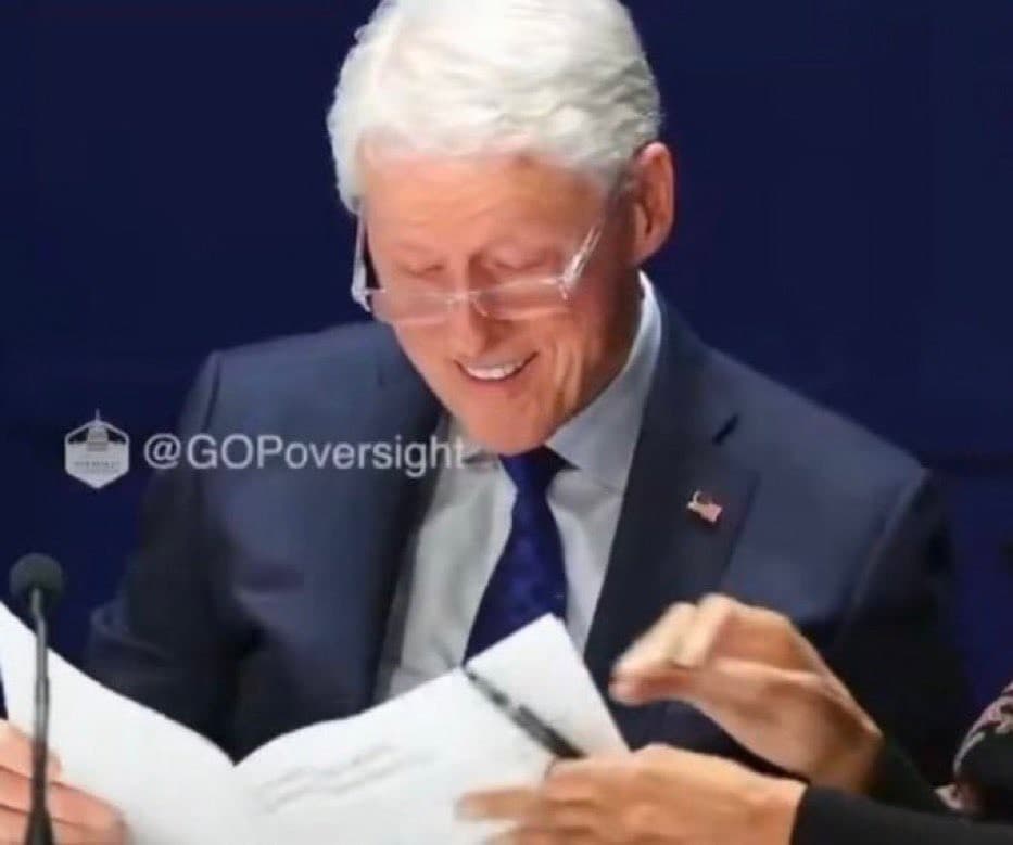 Bill Clinton, wearing a suit and glasses, smiles and laughs while holding and examining several sheets of paper at a desk or hearing table.