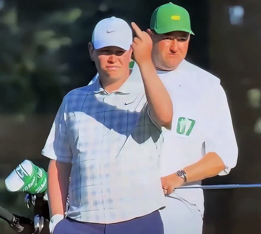 A golfer in a white cap and light shirt stands in front of a caddie wearing a green cap and white bib; the caddie raises one middle finger behind the golfer’s head while both look toward the camera.