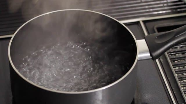 A black pot of water boiling and steaming on a stovetop, with the handle pointing right.