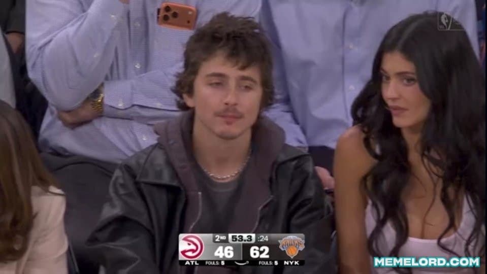 Timothée Chalamet sits courtside in a black leather jacket with a tired, unfocused expression while Kylie Jenner looks toward him; an NBA game scoreboard appears along the bottom of the broadcast image.