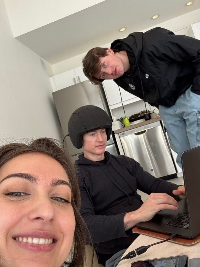 Vertical indoor selfie showing a smiling woman close to the lower-left foreground, a seated man in a black helmet using a laptop near the center, and another man leaning into the frame from the upper-right.