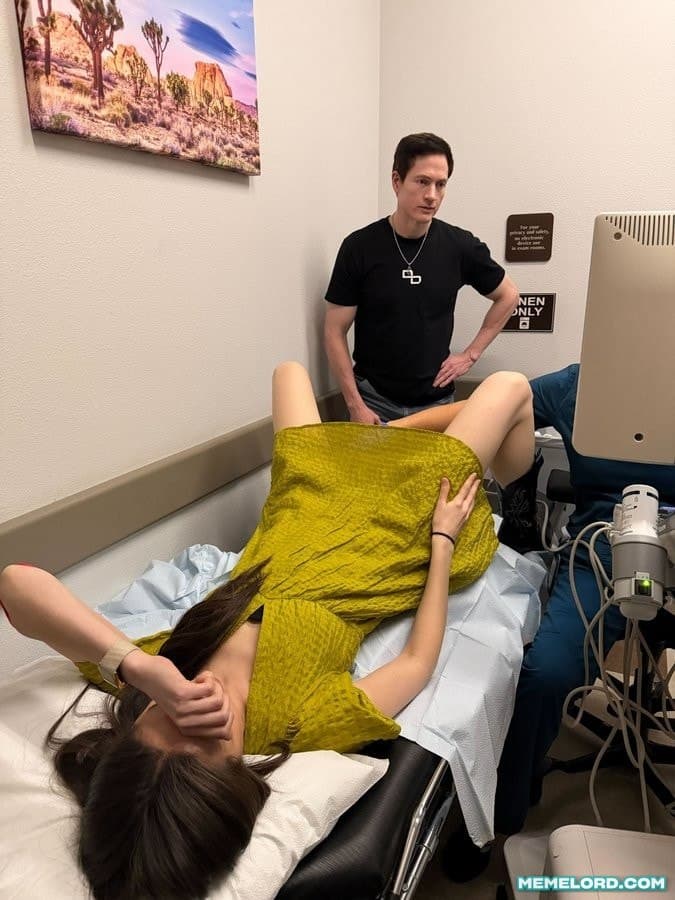 A man in a black shirt stands with his hands on his hips while a woman lies on an examination bed during an ultrasound examination. An ultrasound monitor and medical equipment are visible at the right.