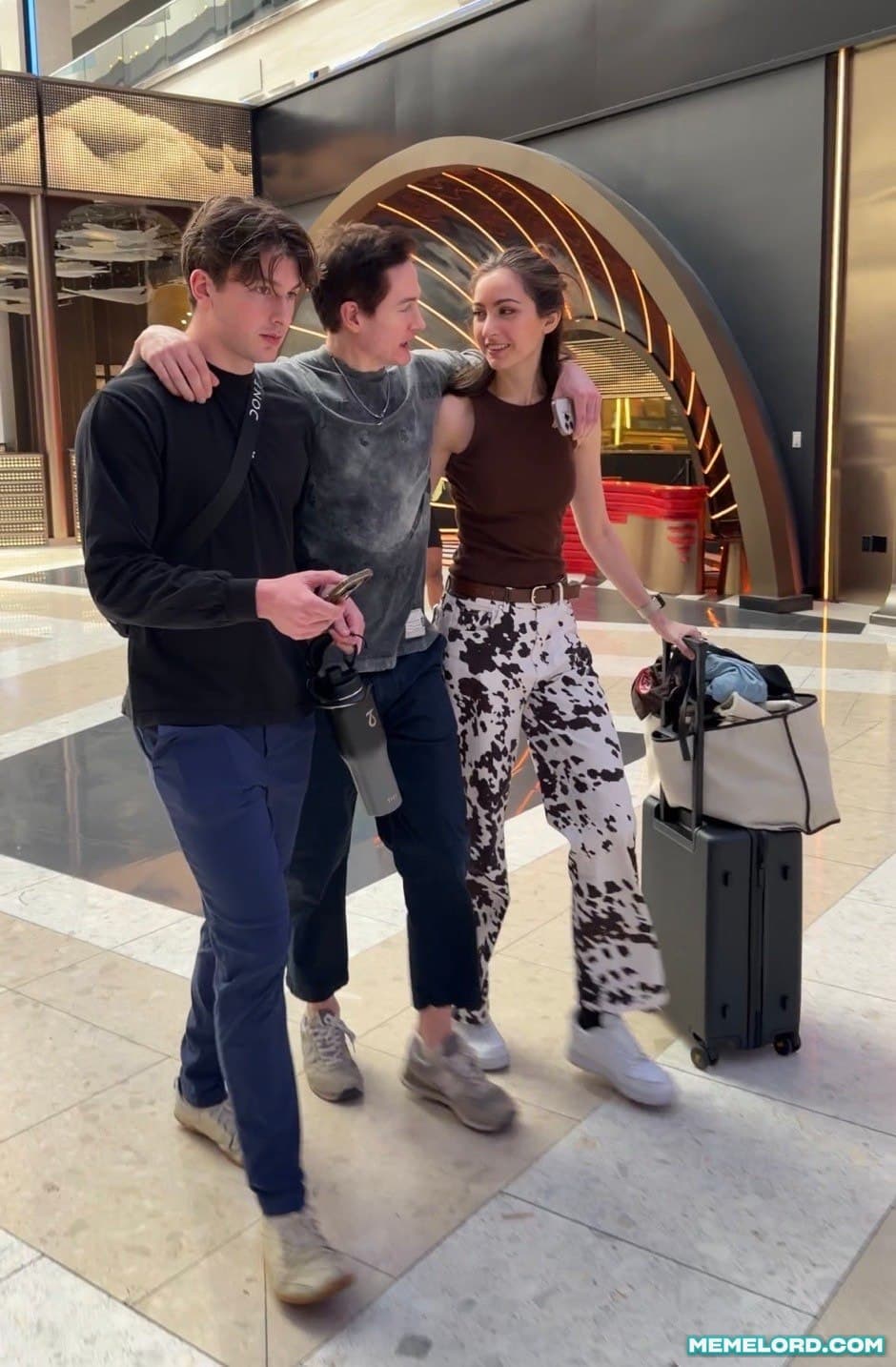 A tall young man, a dark-haired man, and a woman walk together through a brightly lit indoor shopping or airport-like space; the woman pushes a rolling suitcase and a watermark appears at the bottom.