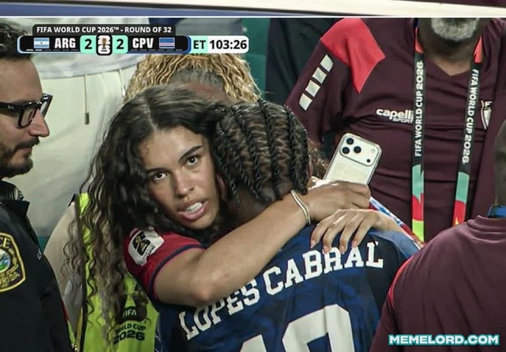 A football player wearing a blue shirt labeled Lopes Cabral embraces a woman with long dark hair while people and a broadcast scoreboard appear around them in a stadium setting.