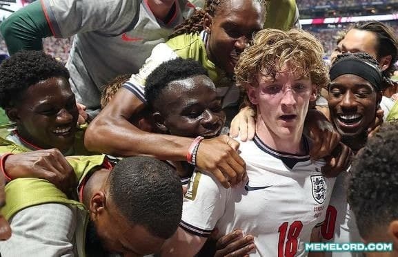 A blond England footballer in a white number 18 jersey is surrounded by several smiling teammates in a close, energetic celebration on a football pitch, with a MEMELORD.COM watermark in the lower-right corner.