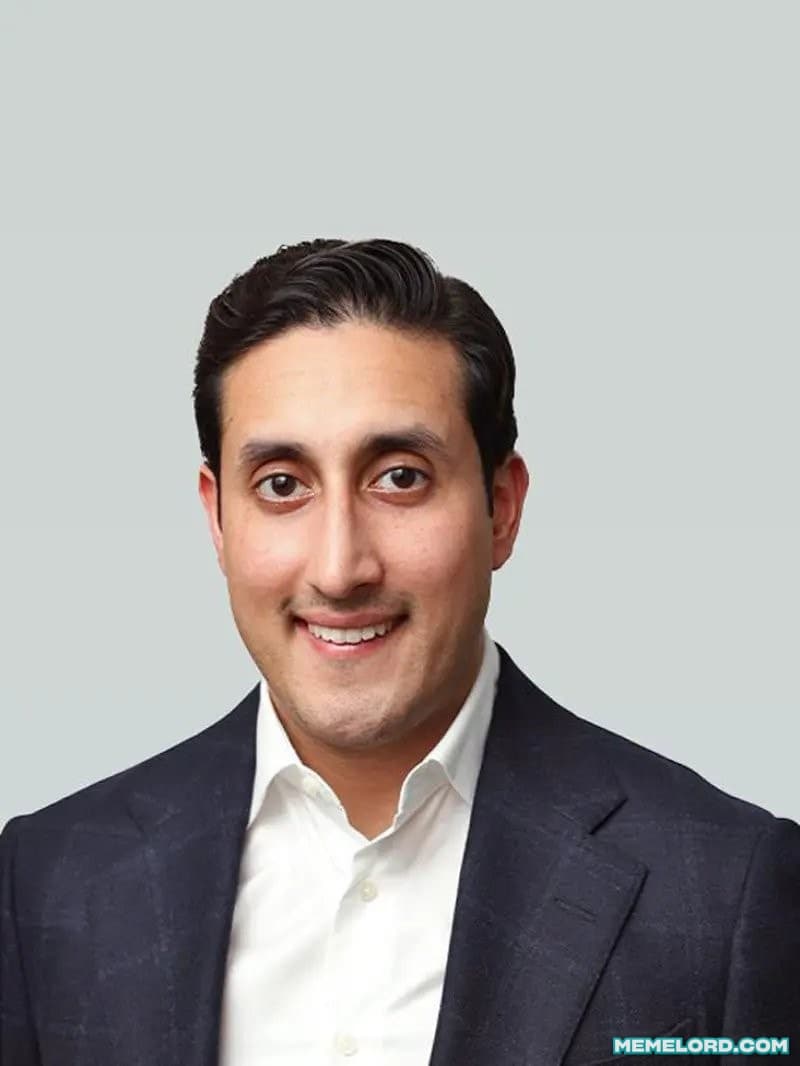 Portrait of a smiling dark-haired man in a navy suit and white shirt against a pale gray background, with a MEMELORD.COM watermark in the lower-right corner.