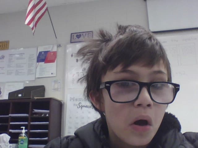 A dark-haired kid wearing large black glasses sits in a classroom with his mouth open and a bewildered expression; a whiteboard, cubbies, and a small American flag are behind him.