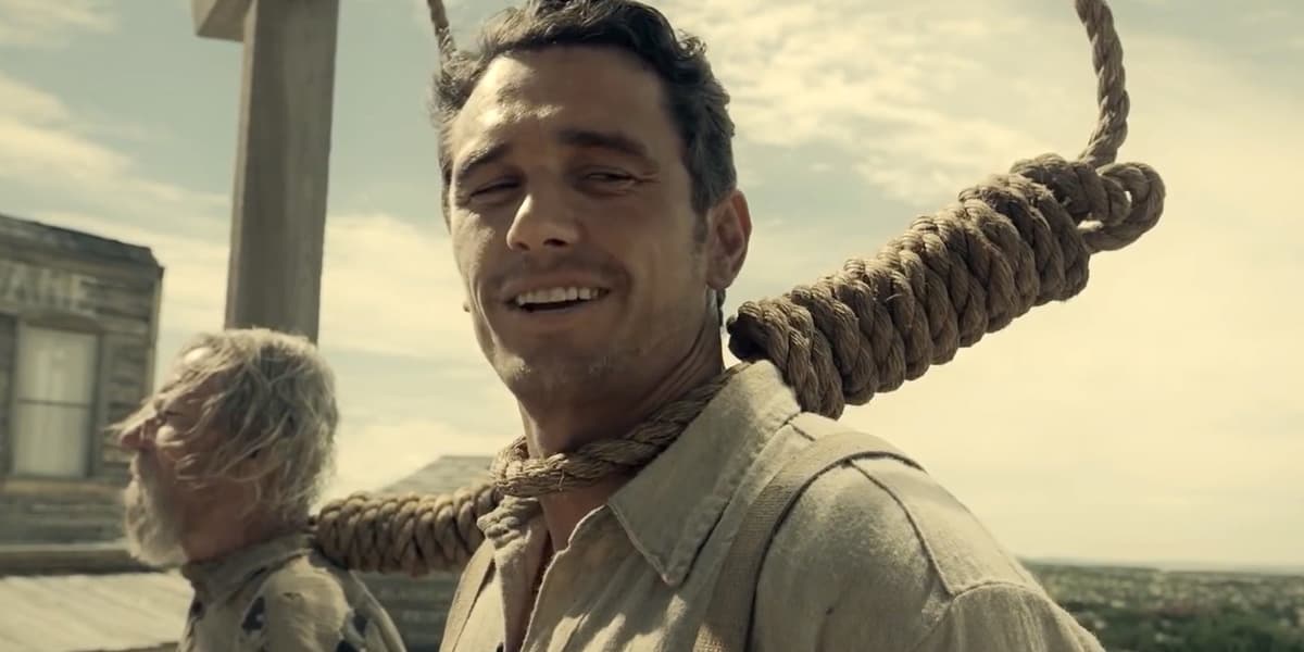 James Franco smiles calmly in period Western clothing while standing with a thick rope noose around his neck; another distressed person is visible behind him near a wooden gallows.