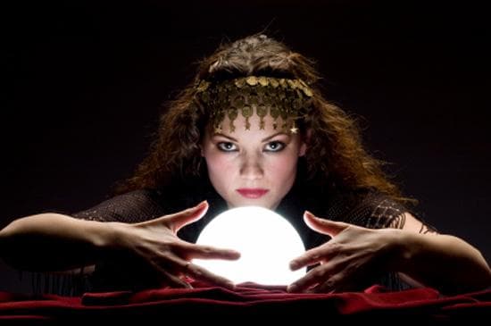 A fortune teller leans over a glowing crystal ball in a dark room, staring intensely toward the viewer.