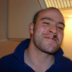 A smiling young man with short hair and a cigarette in his mouth, looking friendly and approachable against a plain background