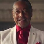 A close-up of actor Giancarlo Esposito smiling warmly at the camera, wearing a white suit jacket over a red dress shirt and red necktie against a warm, blurred interior background.