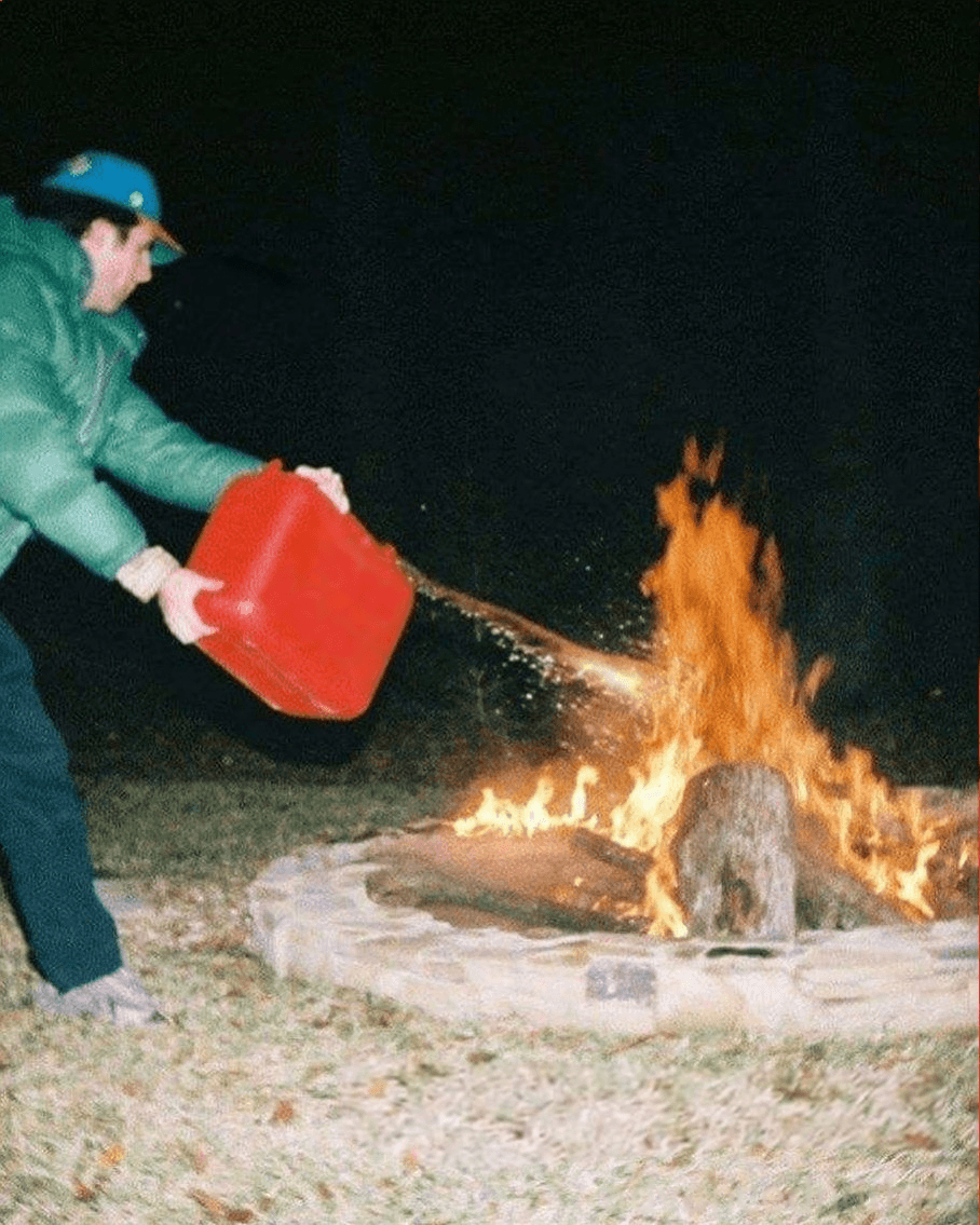 A person in outdoor clothing holding a red gas can and deliberately pouring gasoline onto a campfire, making the flames much larger and more intense. The fire grows dramatically, showing the direct cause-and-effect of making things worse.