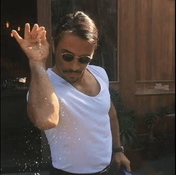 Turkish chef Nusret Gökçe (Salt Bae) in a white t-shirt and sunglasses, dramatically sprinkling salt from above onto meat with his signature arm pose. Salt cascades down in a theatrical fashion.