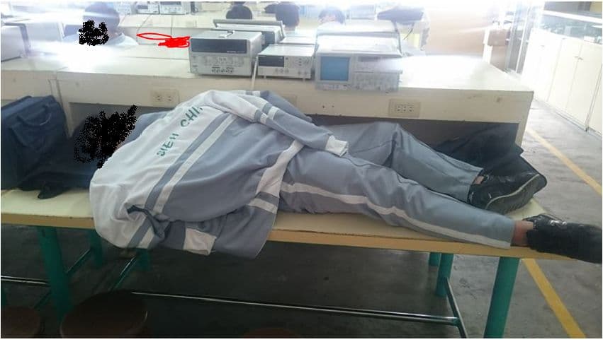 A student in a gray tracksuit lies face-down across a long classroom worktable, with electronics equipment and other students visible behind them.