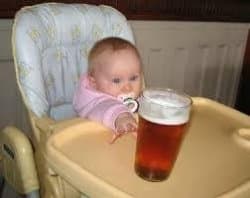 A baby with a pacifier in a high chair reaching one hand toward a full pint glass of amber beer sitting on the tray, just inches away.