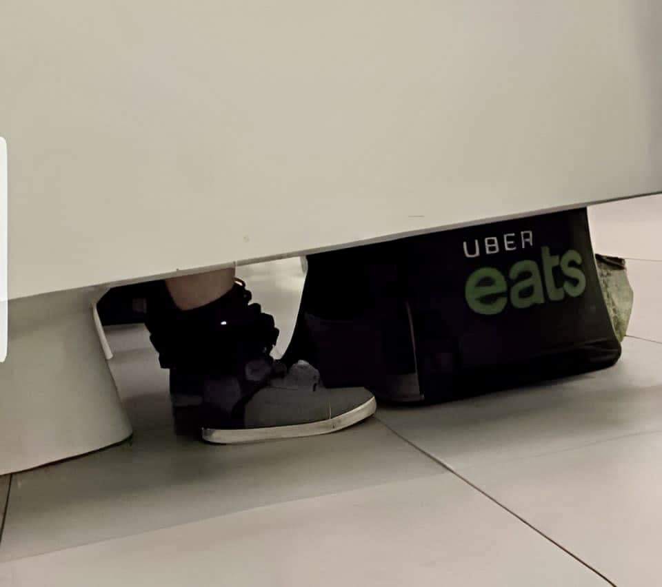 The lower half of a white restroom stall with a person's shoes and an Uber Eats delivery bag visible on the tiled floor beneath it.