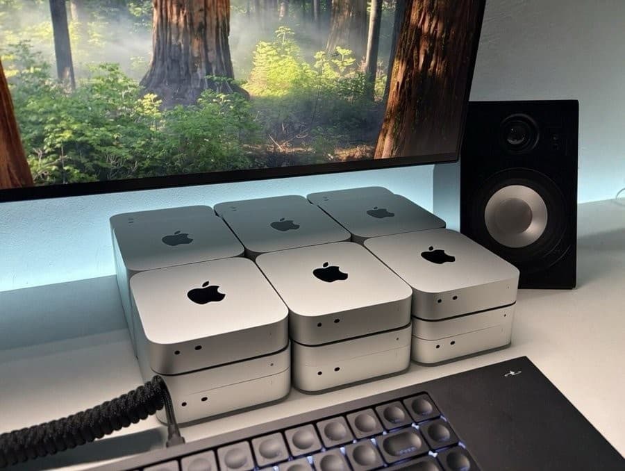 A desktop setup with silver Apple Mac mini computers arranged in several compact stacks beneath a large monitor displaying a sunlit forest. A black studio speaker stands to the right, and part of a keyboard and cable are visible along the bottom edge.