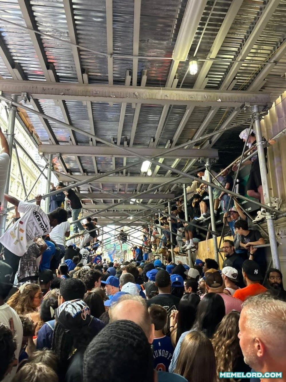 A dense crowd of New York Knicks fans fills a street-level viewing area beneath metal construction scaffolding, while several spectators sit or stand on the horizontal braces and railings above them. Knicks jerseys, caps, and blue clothing are visible throughout the crowd. A MEMELORD.COM watermark appears at the bottom