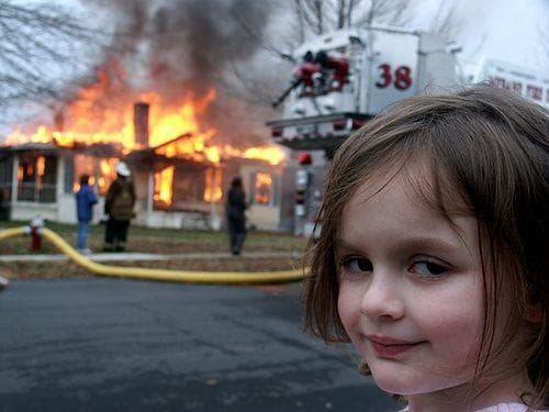 A young girl with brown hair looks and smiles toward the camera in the lower-right foreground while firefighters and a burning house appear behind her on the left.