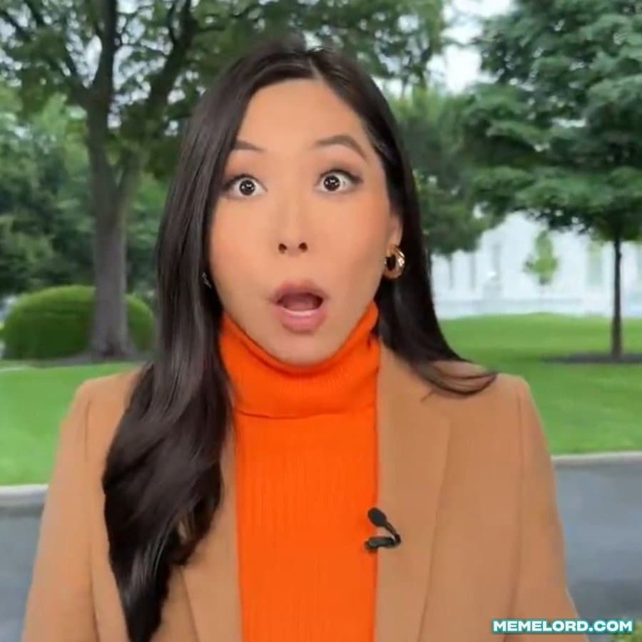 A female reporter in a tan coat and bright orange turtleneck stares directly at the camera with wide eyes and an open mouth, standing outdoors near a white building and trees.