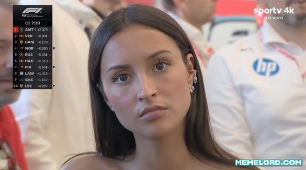 A woman with long dark hair stares directly at the camera with a flat, unimpressed expression during a televised Formula 1 event; an F1 timing graphic appears at upper left and broadcast watermarks are visible.