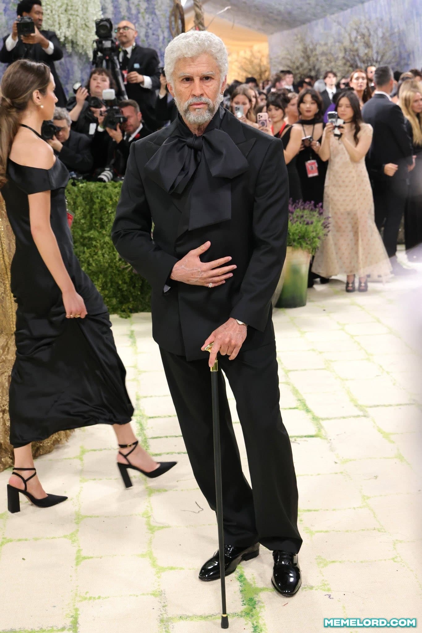 A silver-haired, bearded man in an oversized black formal suit with a large bow at the neck stands in the center of a crowded gala while holding a cane; photographers and fashionably dressed guests surround him, and a woman in a black dress stands at left.