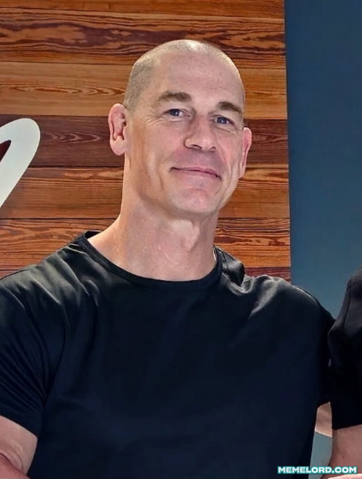 John Cena, appearing bald and wearing a black shirt, looks toward the camera with a slight smirk in front of a wooden wall.