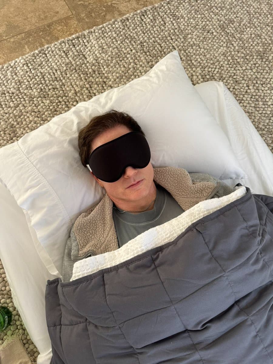 A man wearing a black sleep mask lies on a white pillow under a gray blanket, photographed from above.