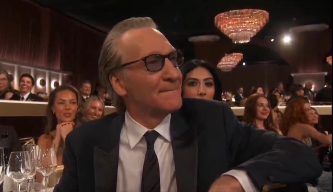 Bill Maher wearing dark glasses and a suit sits at a formal event, looking to the side with a tight-lipped, amused expression while an audience watches behind him.