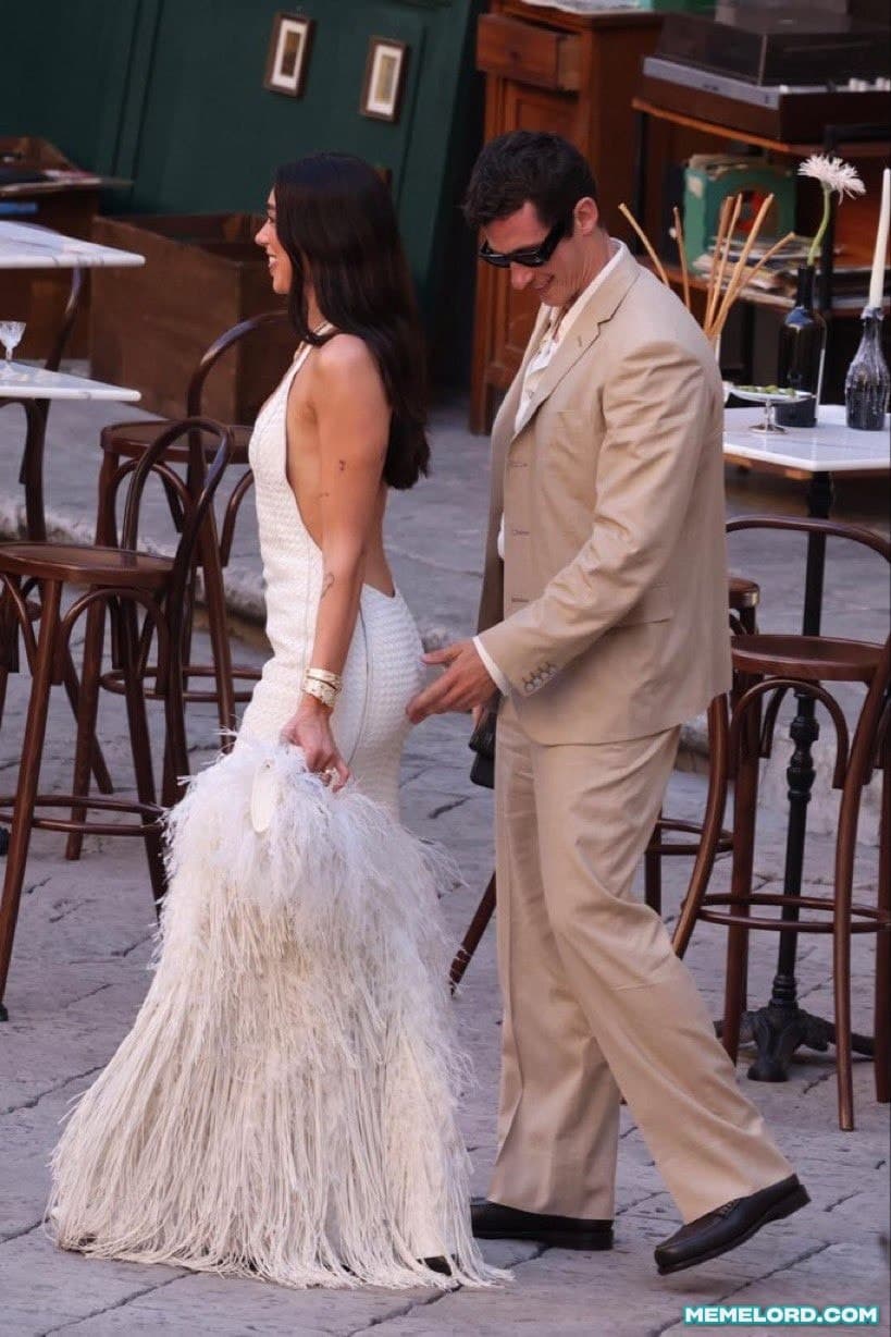 A man in a cream suit and dark sunglasses walks behind a woman in a white feathered dress while looking downward toward her.