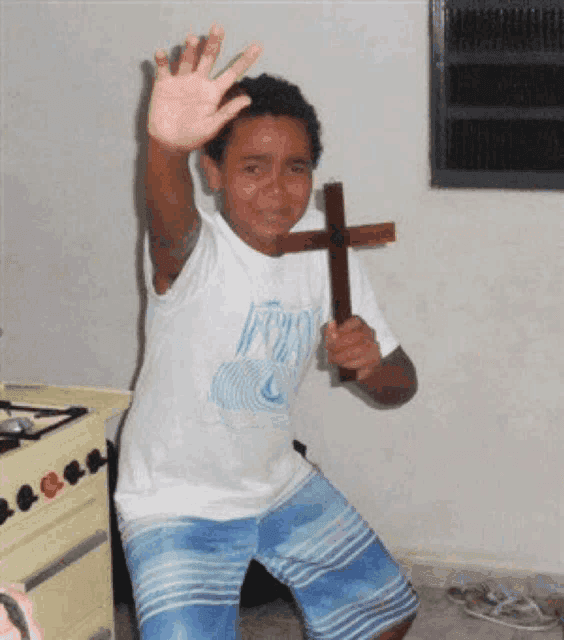 A young boy with curly hair cries in fear while holding a small wooden cross in one hand and raising his other palm outward in a stop gesture, standing in a kitchen near a stove.