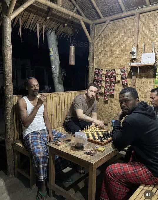Snoop Dogg, Eminem, and 50 Cent sitting together in a small bamboo hut